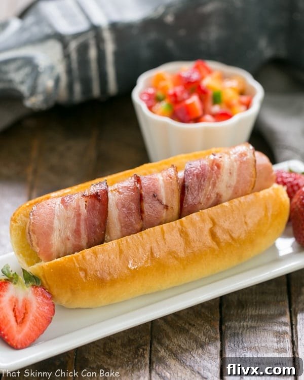 Two perfectly cooked Bacon Wrapped Hot Dogs adorned with fresh Fruit Salsa on a sleek rectangular plate, accompanied by a small bowl of extra salsa.