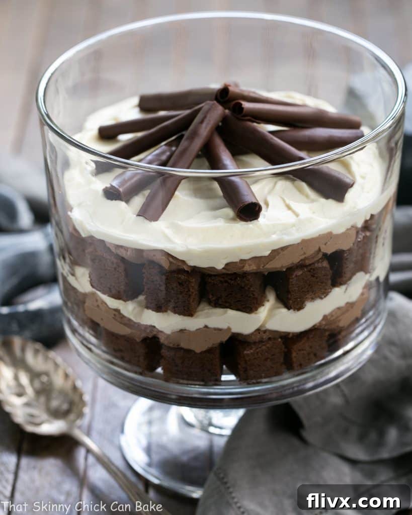 A beautifully layered Fudgy Brownie Trifle with Chocolate Mousse in a clear glass trifle dish, ready to be served with a silver spoon. Top is garnished with chocolate curls.