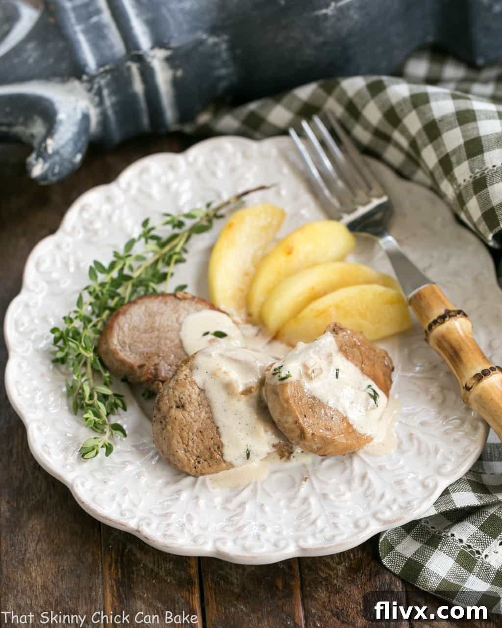 Apple Topped Pork Medallions with Calvados Cream Sauce served on a dinner plate with fresh thyme garnish and a bamboo handle fork, ready to eat.