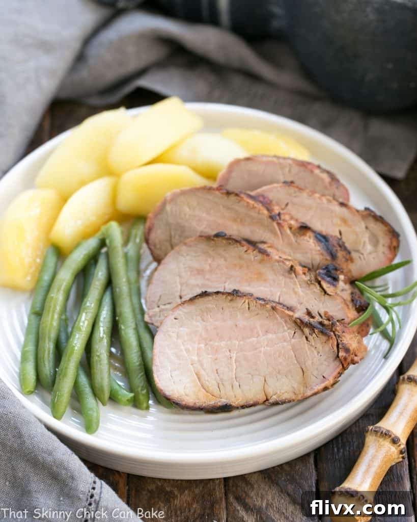 Close-up of perfectly sliced Maple Grilled Pork Tenderloin medallions, garnished with fresh rosemary, served alongside vibrant green beans and caramelized sauteed apples on a white plate.
