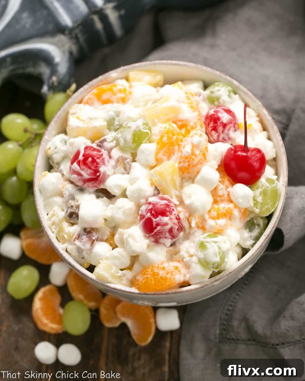 Ambrosia Salad in a white ceramic bowl surrounded by fresh grapes, mini marshmallows and mandarin orange segments.