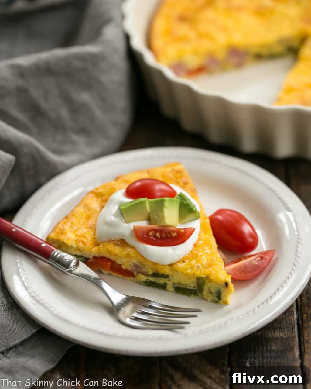 A vibrant slice of Baked Denver Omelet on a white plate, artfully garnished with fresh avocado and cherry tomatoes, with the casserole dish subtly blurred in the background, hinting at more deliciousness.
