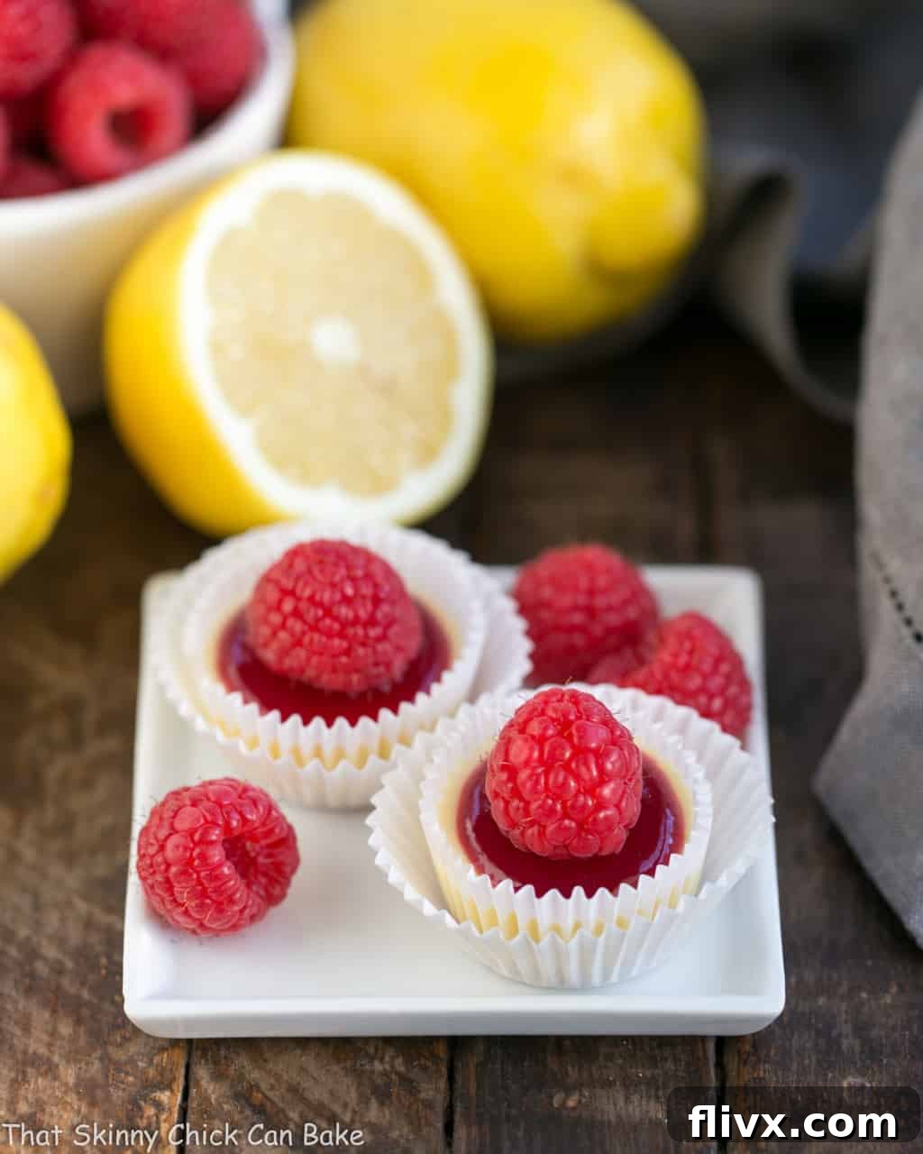 Two exquisite raspberry-topped cheesecake bites presented on a small white plate, adorned with additional fresh raspberry garnishes, highlighting their elegant appeal.