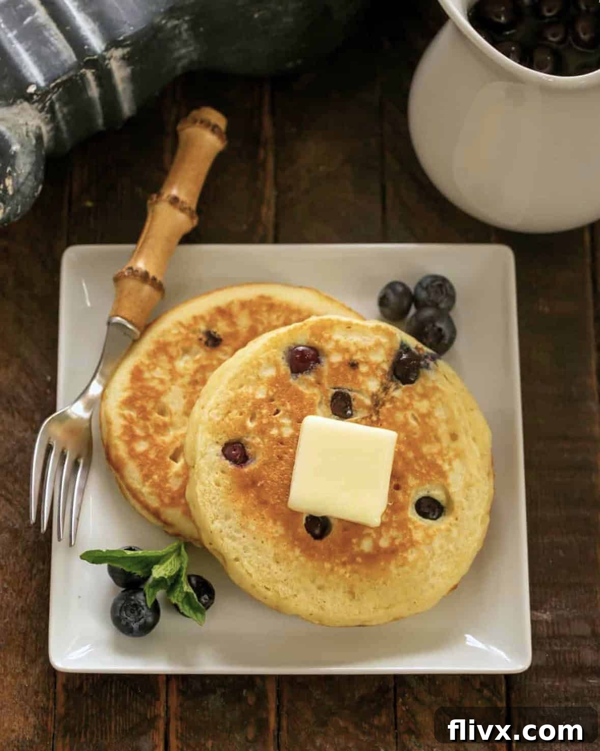 Two fluffy blueberry buttermilk pancakes on a square white plate, with a stylish bamboo handle fork resting beside them.