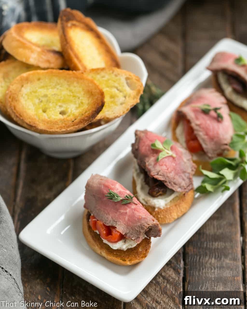 Close-up overhead view of Beef Crostini with Blue Cheese Spread artfully arranged on a narrow white tray, highlighting the fresh ingredients and elegant presentation.