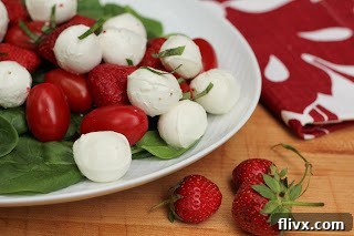 A beautifully arranged oval platter featuring a vibrant Mozzarella, Tomato, and Strawberry Salad, showcasing fresh bocconcini, bright red grape tomatoes, juicy sliced strawberries, and a sprinkle of green basil leaves, all glistening with a balsamic vinaigrette.