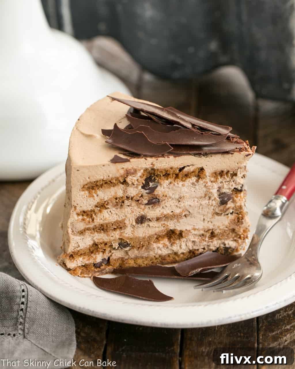 A perfectly sliced piece of No-Bake Mocha Chocolate Chip Cookie Icebox Cake on a pristine white dessert plate, accompanied by a festive red-handled fork, ready to be enjoyed. Layers of creamy mocha and crisp chocolate chip cookies are visible.