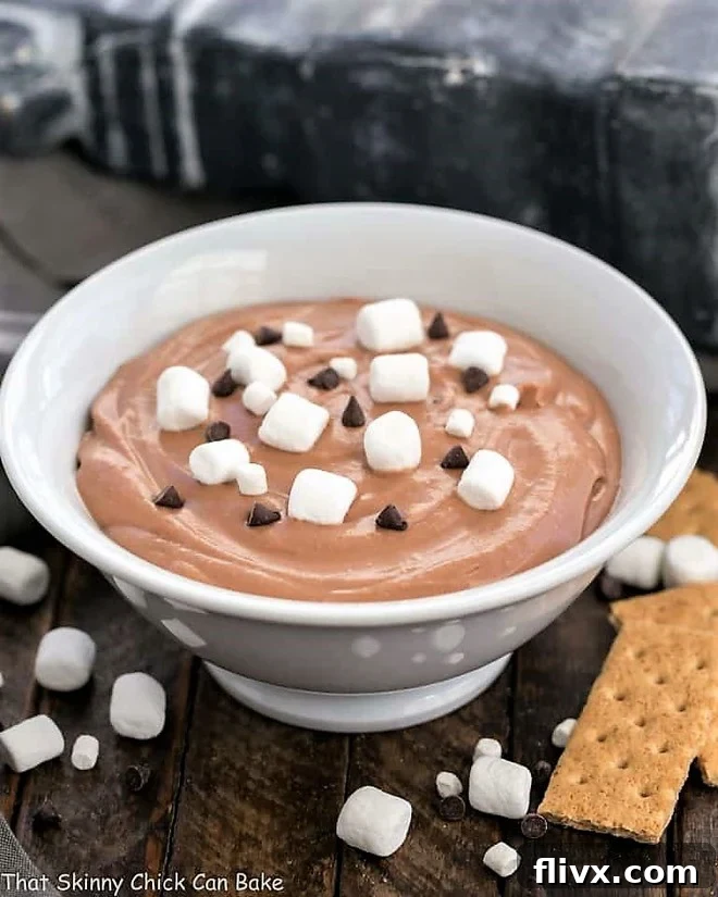 A close-up shot of the finished Easy S'mores Dessert Dip in a white ceramic bowl, beautifully garnished with a generous scattering of fluffy mini marshmallows and rich chocolate chips, looking absolutely ready to be enjoyed.
