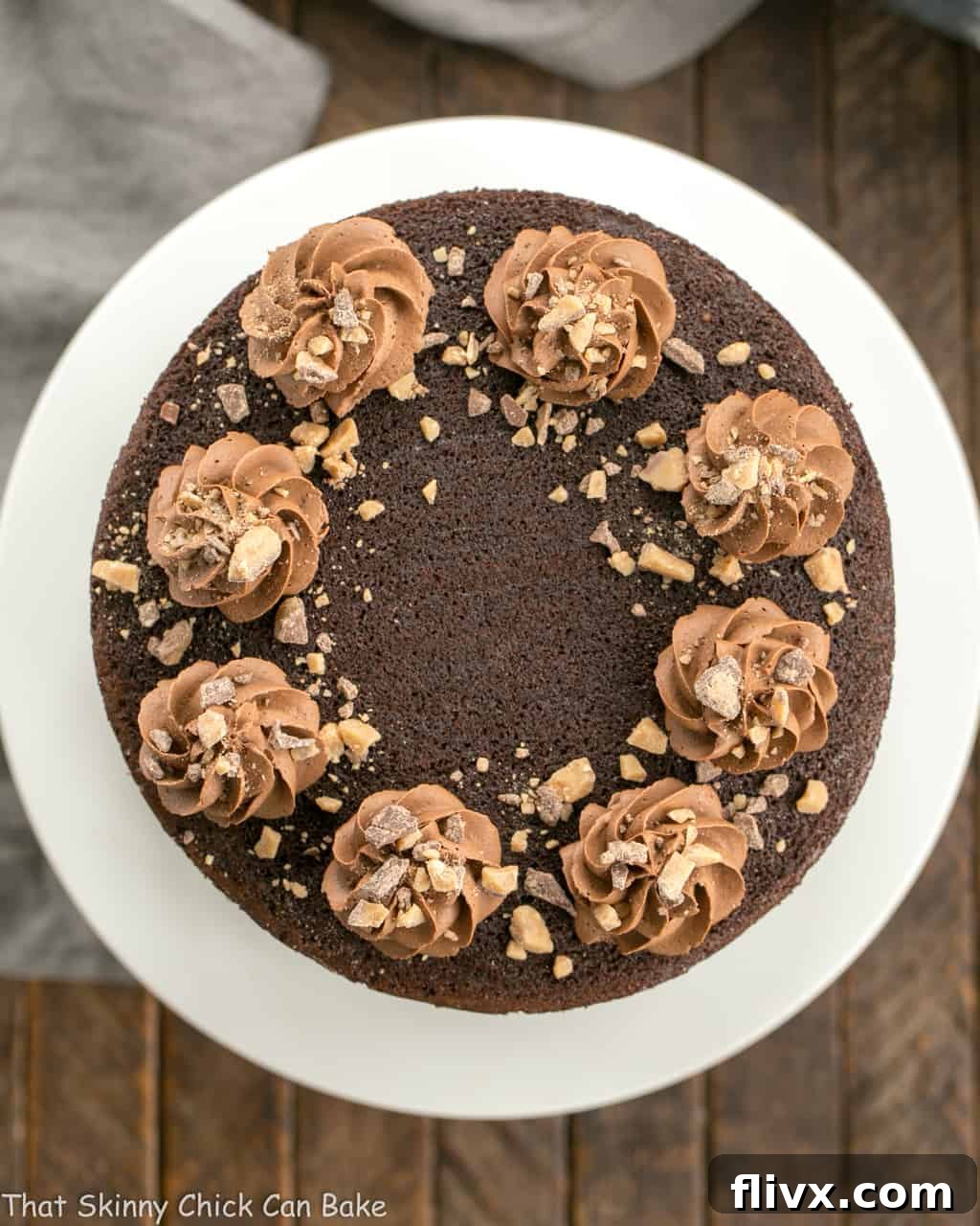 Overhead view of a Triple Layer Chocolate Toffee Cake on a white cake stand, garnished with piped frosting swirls and toffee bits.