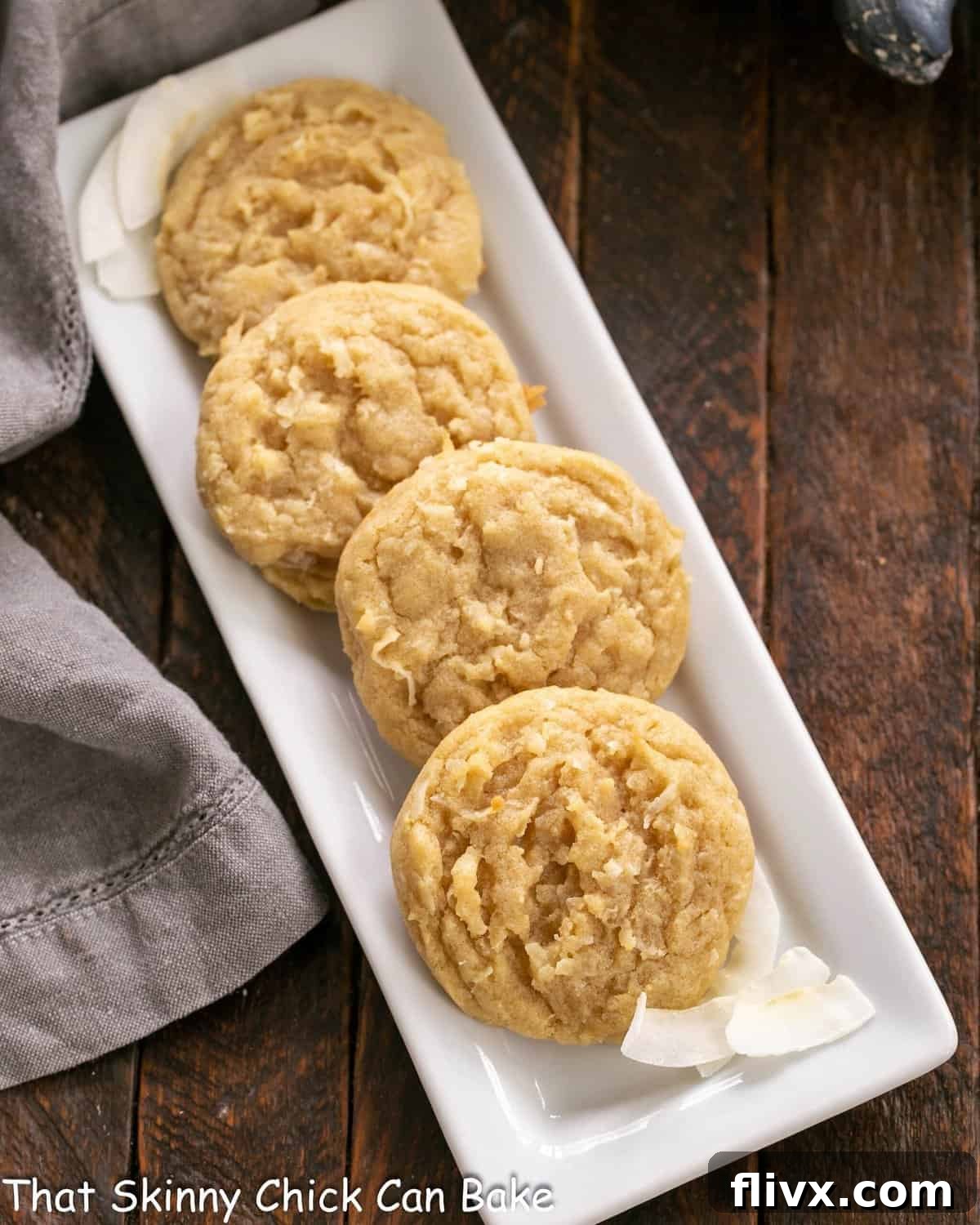 Overhead view of 4 perfectly baked chewy coconut cookies on a pristine white ceramic tray, showcasing their golden-brown edges and moist, shredded coconut texture.