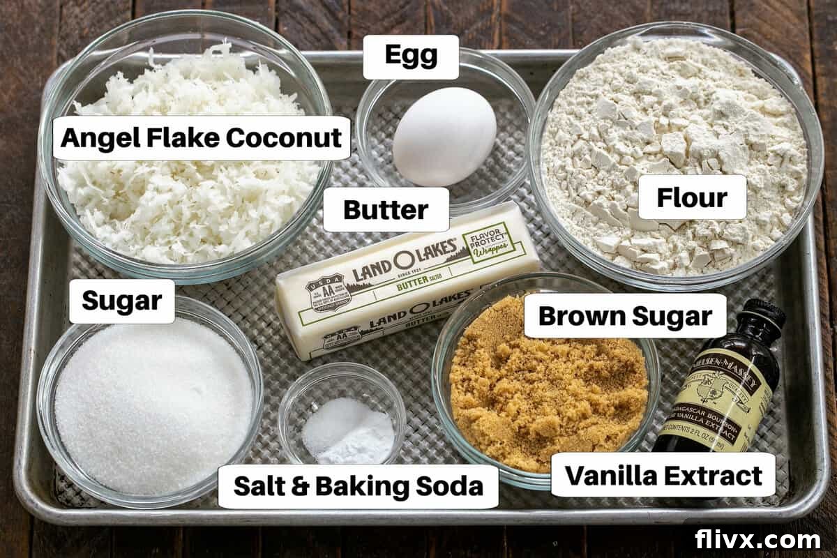 All the essential ingredients for making chewy coconut cookies laid out on a sheet pan, clearly labeled for easy identification: butter, brown sugar, granulated sugar, egg, vanilla extract, flour, baking soda, salt, and sweetened shredded coconut.