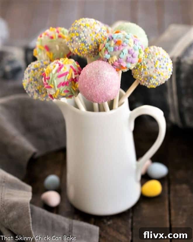 A cluster of beautifully decorated Easter Cake Pops, displayed upright in a rustic ceramic pitcher, showcasing pastel sprinkles against a white chocolate coating.