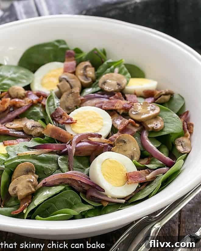 Best Spinach Salad close up in a white ceramic bowl. Fresh spinach, crispy bacon, hard-boiled eggs, and sautéed mushrooms and onions are visible, drizzled with a rich bacon dressing.