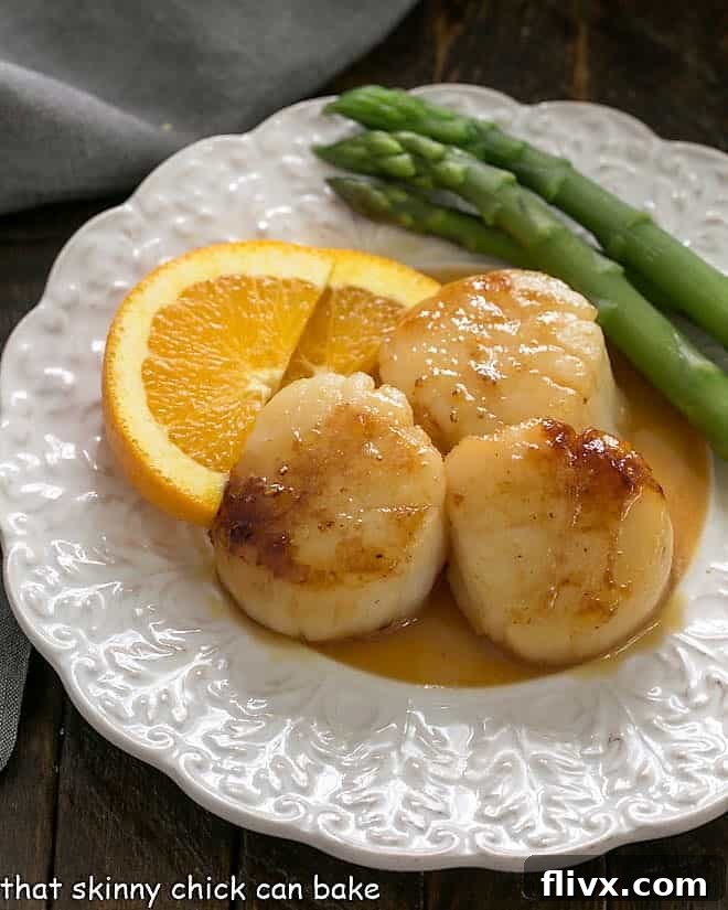 Pan seared scallops on a decorative white plate with orange slices and asparagus, highlighting the rich sauce.