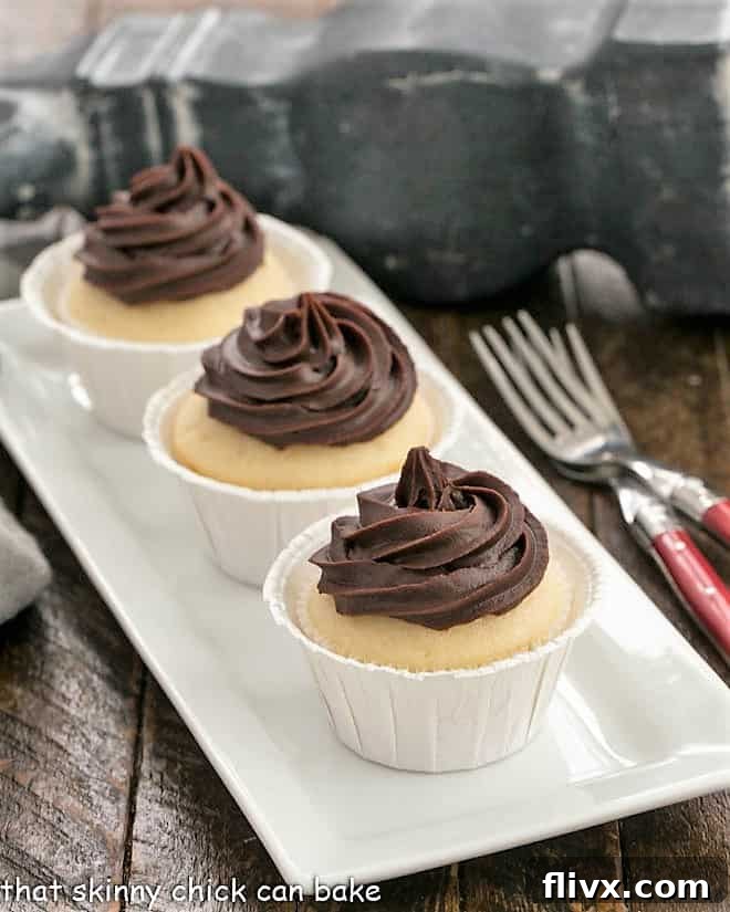 Three Boston cream pie cupcakes arranged on a pristine white tray, accompanied by two elegant red-handled forks, highlighting their inviting appearance and readiness for serving.