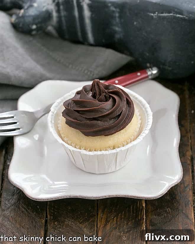 A single Boston Cream Pie Cupcake artfully placed on a small, elegant white plate, showcasing its golden cupcake base, a hint of creamy filling, and a rich swirl of chocolate ganache on top.