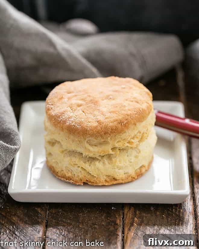 A golden-brown buttermilk biscuit resting on a square white plate, accompanied by a red-handled knife.