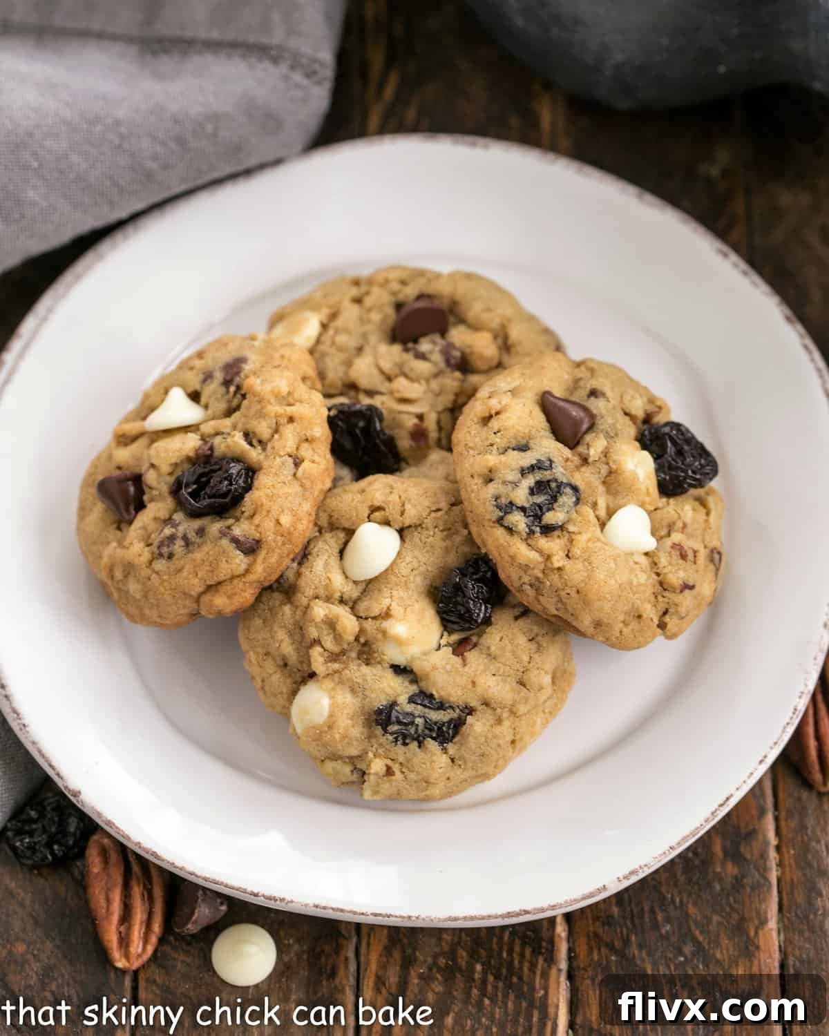 An overhead view of freshly baked chewy oatmeal cookies, adorned with generous chocolate chunks, pecans, and dried cherries, artfully arranged on a pristine white plate.