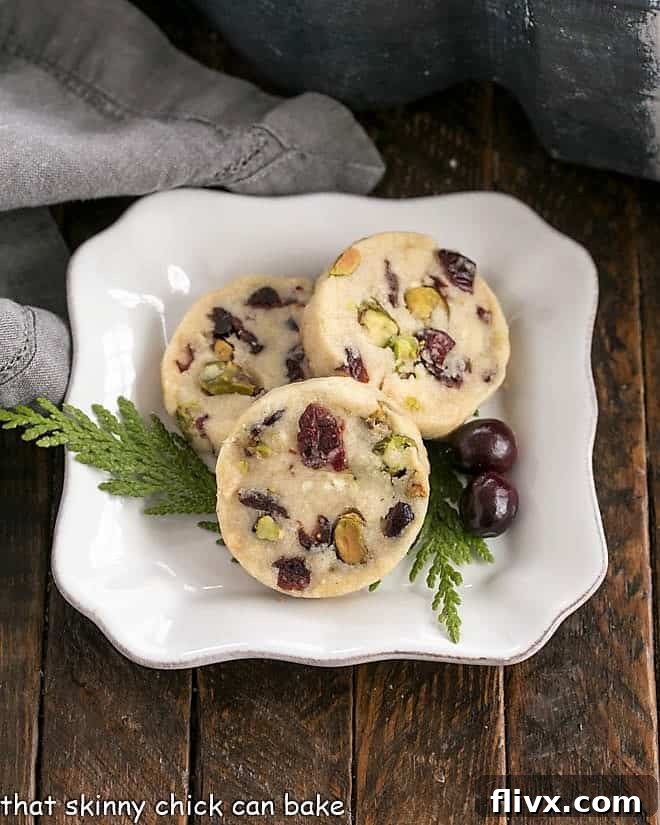 Three golden brown Cranberry Pistachio White Chocolate Shortbread Cookies on a square white plate, with a subtle festive background.