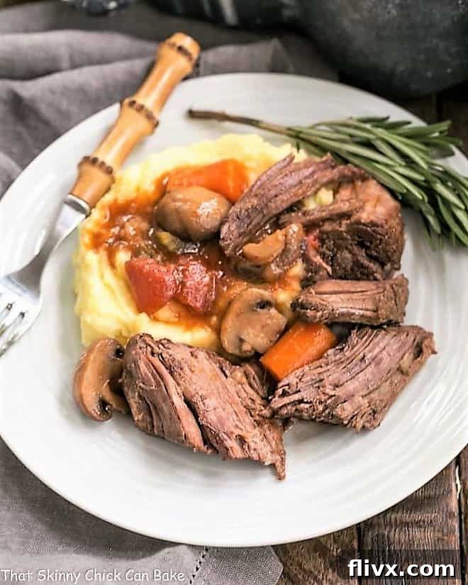 A generous serving of Oven Pot Roast with Mushrooms, Tomatoes & Red Wine on a white plate, accompanied by creamy mashed potatoes and a fresh sprig of rosemary.