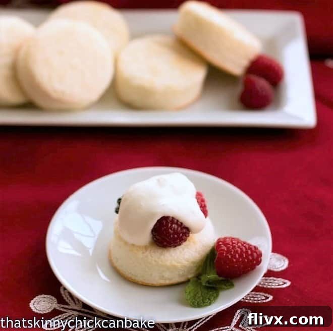 A close-up view of an individual Emeril's Angel Cake Sandwich, with the top cake layer removed to reveal a generous filling of fresh raspberries and whipped cream, garnished with a single raspberry on a pristine white plate.