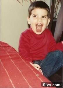 The birthday boy, Tom Berg, as a child in a red shirt and jeans, smiling broadly, ready to eat his chocolate cake.