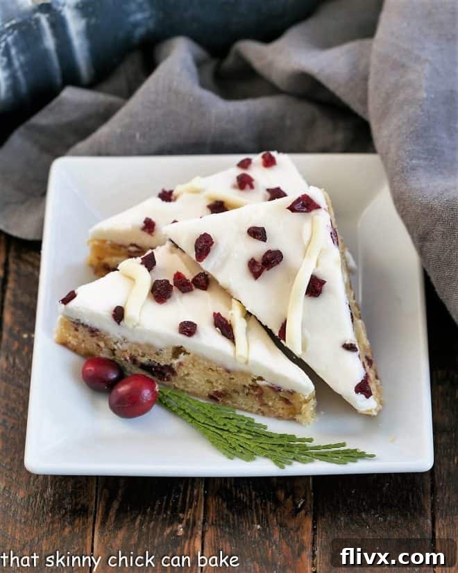 Three cranberry bliss bars elegantly arranged on a square white plate, garnished with fresh cranberries and a sprig of cedar.