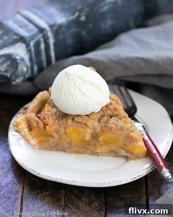 A delectable slice of Easy Peach Crumb Pie, crowned with a scoop of vanilla ice cream, presented on a round white plate. The full pie is visible in the background.