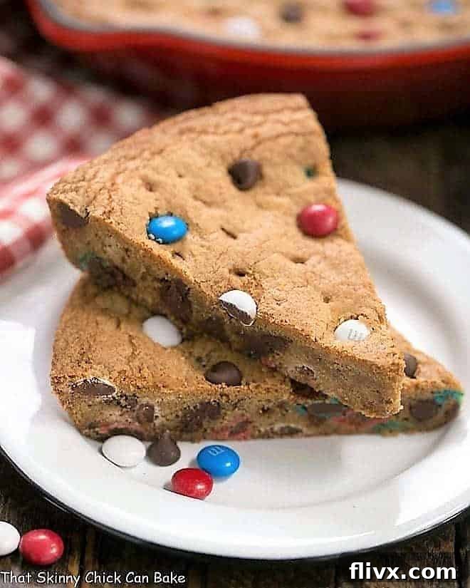 A stack of warm, gooey Patriotic Skillet Cookie wedges on a pristine white enamel dessert plate, showing off melted chocolate and colorful M&M's.