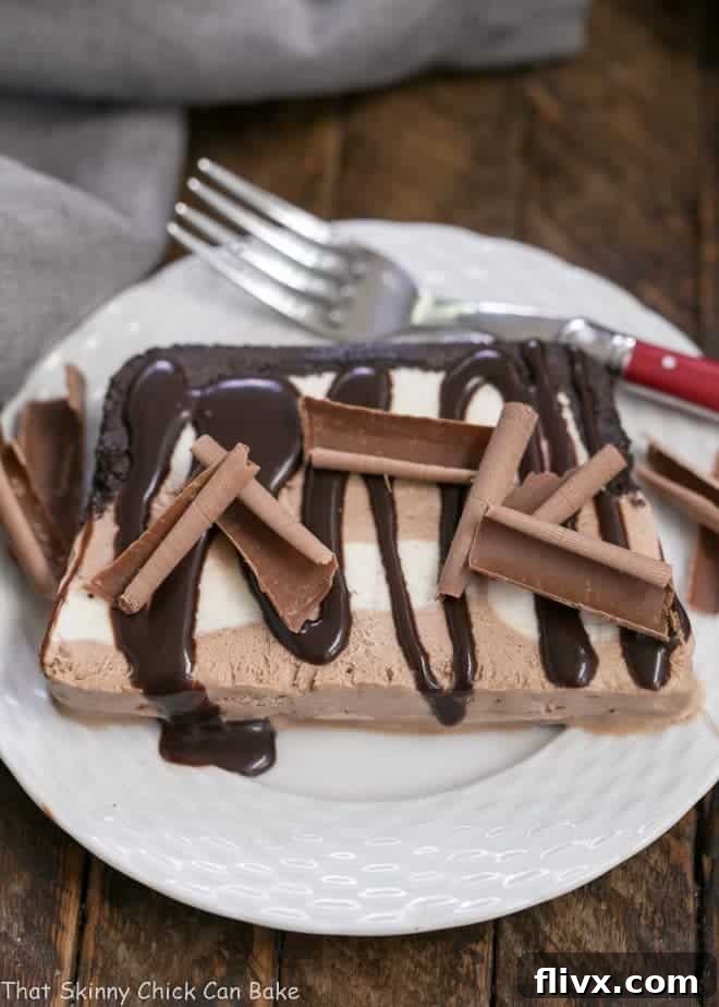 A pristine slice of Homemade No-Churn Ice Cream Cake on a white plate, garnished with chocolate curls and a drizzle of fudge sauce, ready to be enjoyed with a red-handled fork.