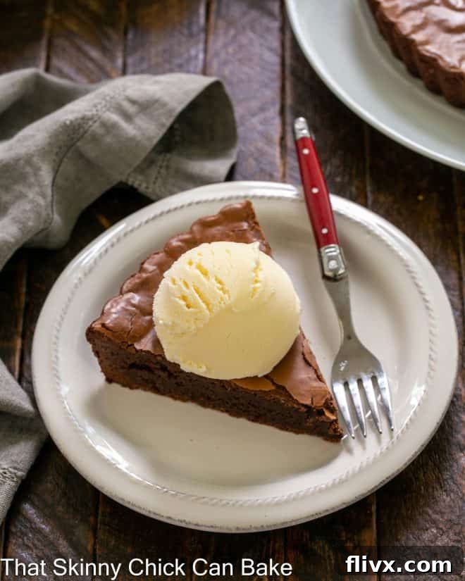 A slice of brownie tart topped with a scoop of vanilla ice cream on a round white plate with a red handled fork.