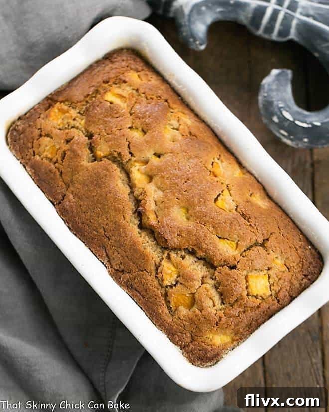 A freshly baked loaf of Southern Peach Bread sits in a white ceramic loaf pan, cooling. The golden-brown crust hints at its rich flavor.