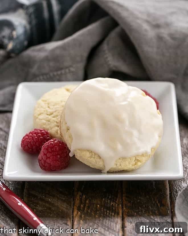 Two super moist Vanilla Bean Scones, delicately glazed and served on a square white plate with a scattering of fresh raspberries, inviting a delightful taste experience.
