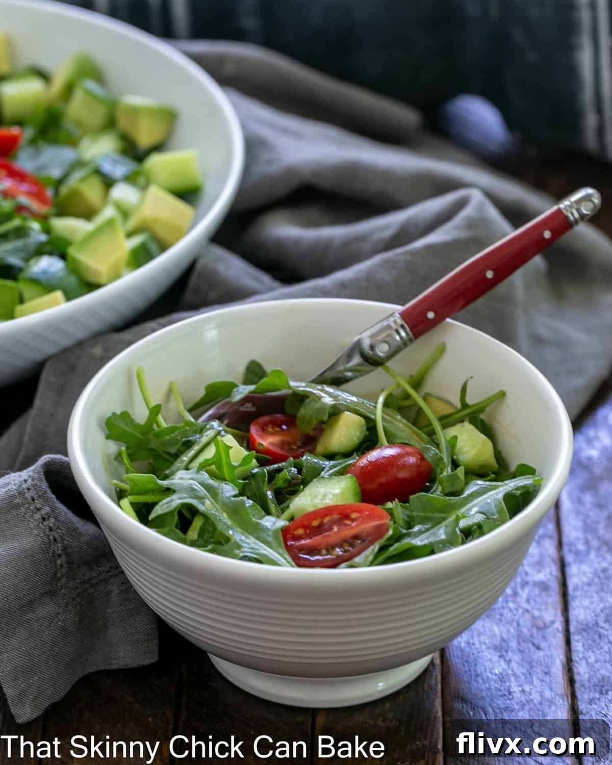 Bright and Peppery Arugula Salad 3 Close-up of a serving bowl filled with fresh Arugula Salad, garnished with halved cherry tomatoes and sliced cucumbers, with a red-handled fork resting in front.