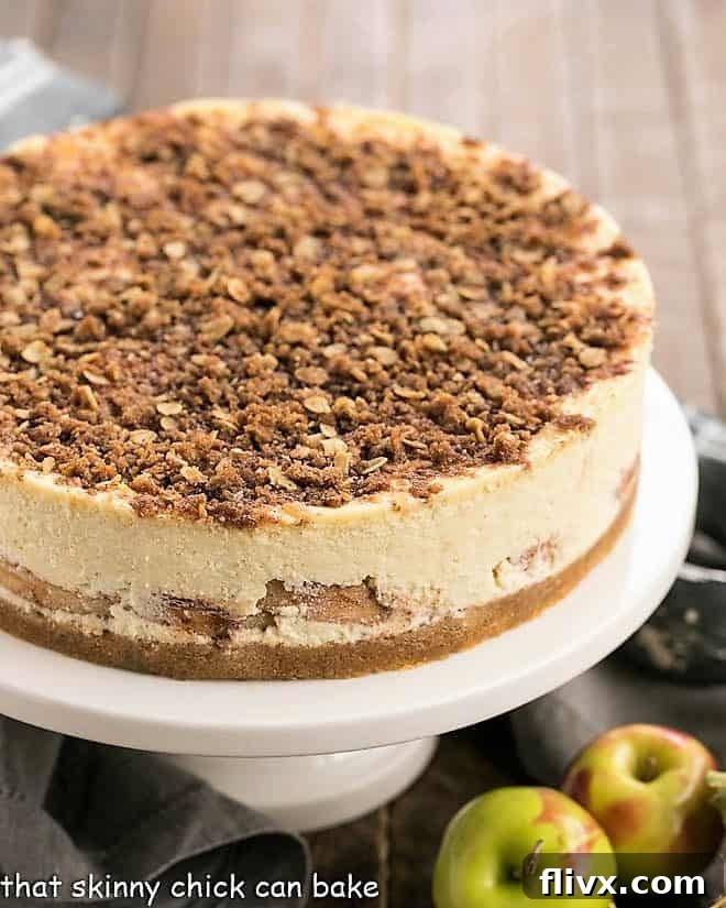 A stunning whole Apple Crisp Cheesecake sitting majestically on a white cake stand, ready to be served. The golden streusel is perfectly crumbled over the smooth cheesecake layer, and a glimpse of the apple filling is visible at the edges.