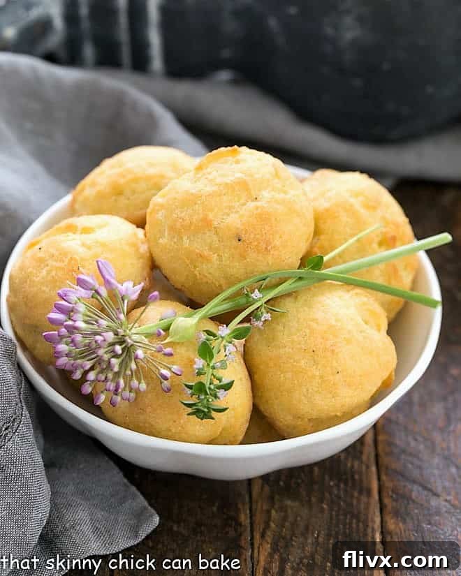 Golden-brown Gougères in a white ceramic bowl, garnished with fresh herbs like chives and thyme, ready to be served as an elegant appetizer.