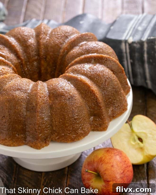 A stunning Apple Bundt Cake sitting elegantly on a white cake stand, flanked by two fresh, vibrant red apples, highlighting its natural goodness and perfect autumn appeal.