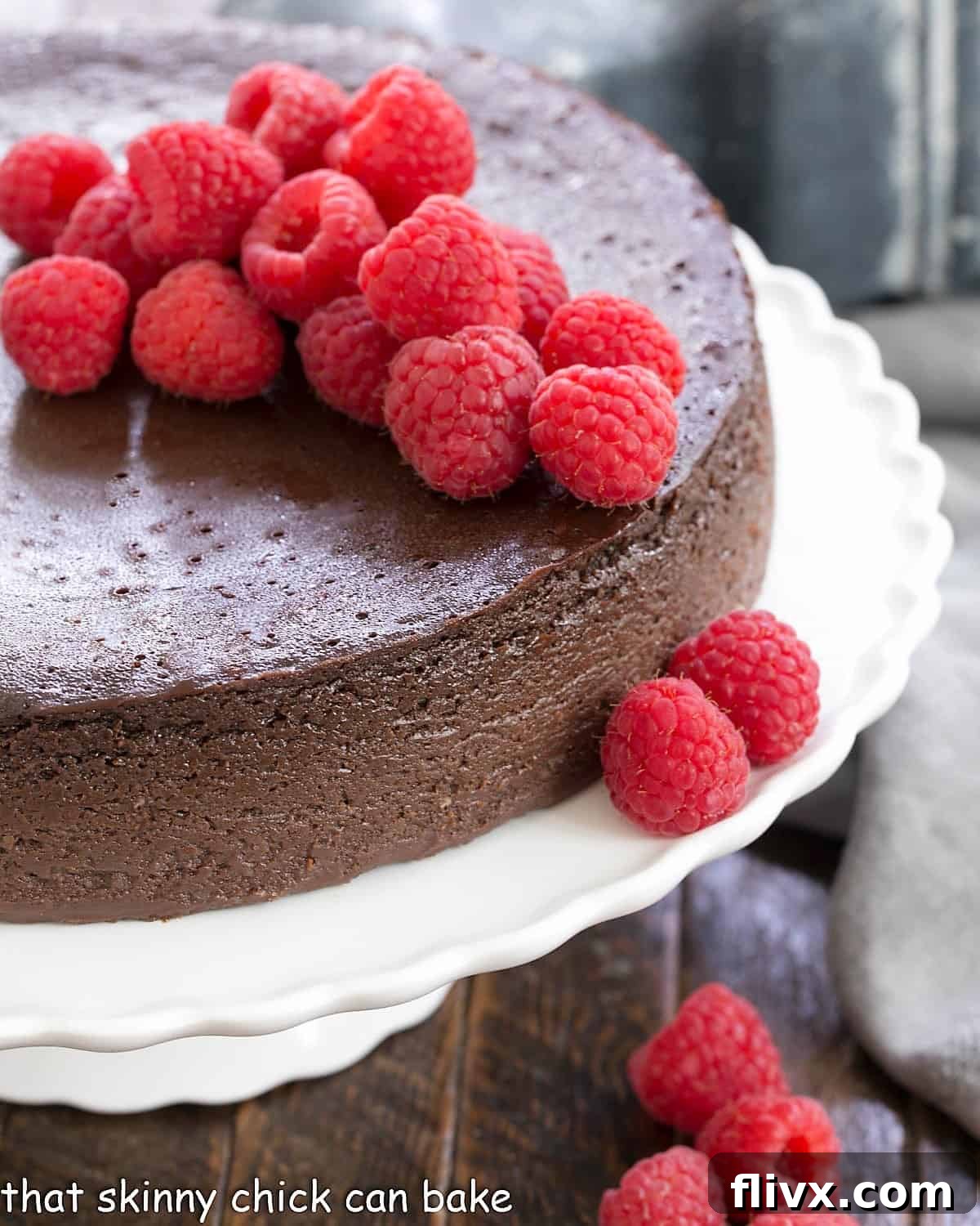 A stunning Flourless Chocolate Kahlua Cake, generously topped with an abundance of vibrant fresh raspberries, presented on a pristine white cake stand.