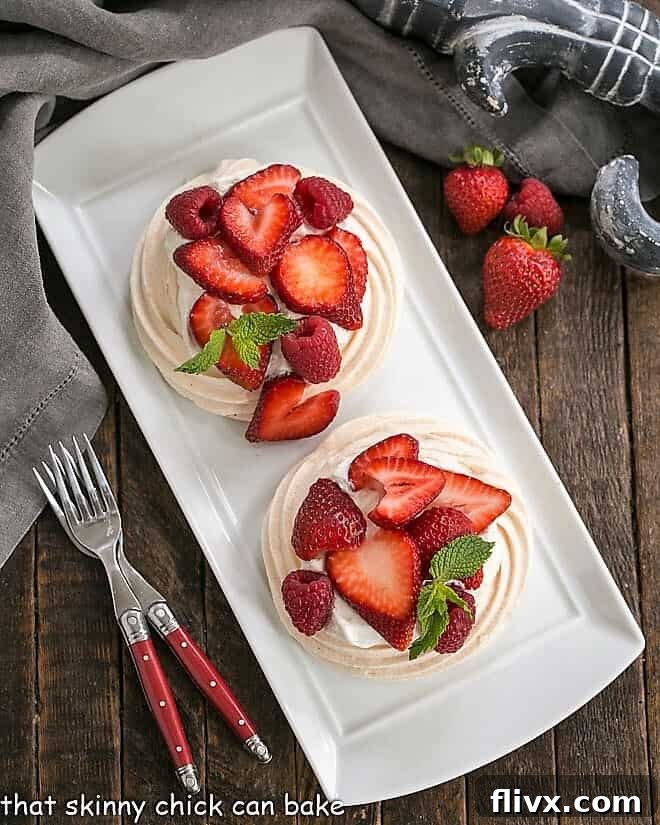 Overhead perspective of two elegantly garnished meringue desserts resting on a white serving tray, accompanied by two red-handled forks