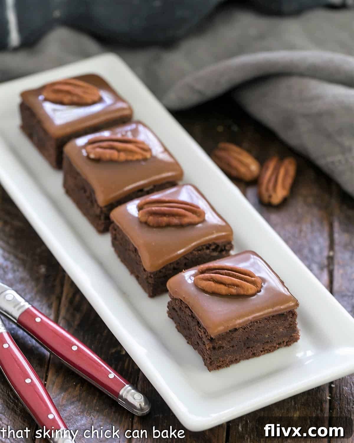 A white tray filled with perfectly cut caramel brownies, garnished with pecan halves, with two red-handled forks poised for enjoyment.