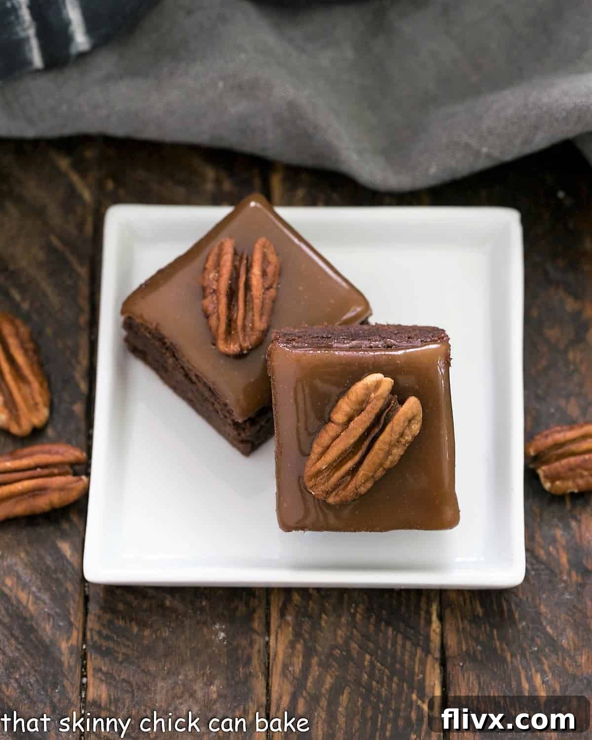 Two perfectly cut turtle brownies, each adorned with a glossy caramel swirl and a whole pecan half, resting on a small square white plate.