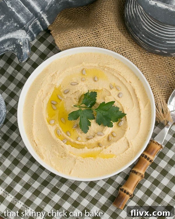 An overhead view of luscious Classic Hummus presented in a white ceramic bowl, elegantly finished with a generous swirl of extra virgin olive oil, a sprinkle of toasted seeds, and a flourish of fresh parsley for garnish.