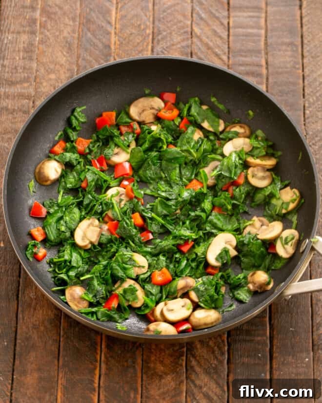 Spinach and diced red bell pepper added to sautéed mushrooms in a skillet, wilting.
