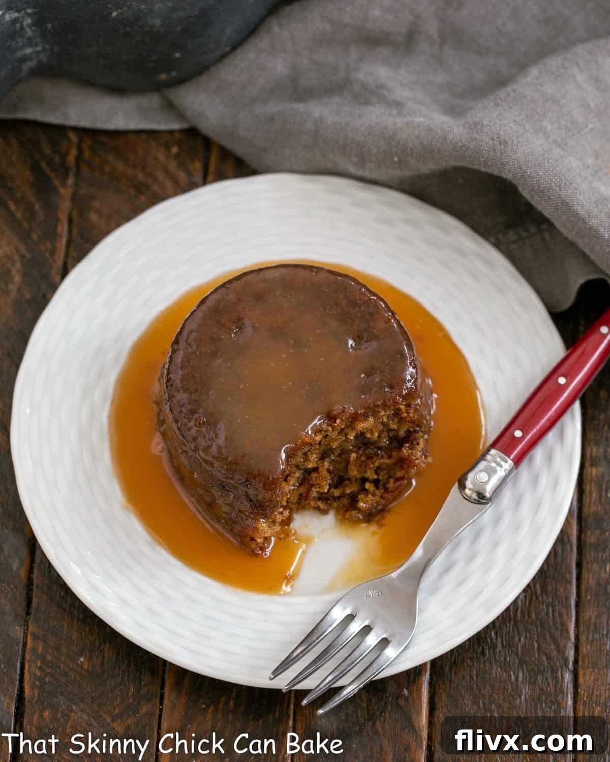 An overhead shot of a sticky toffee pudding served on a pristine white dessert plate, with a red-handled fork resting beside it, inviting a bite.