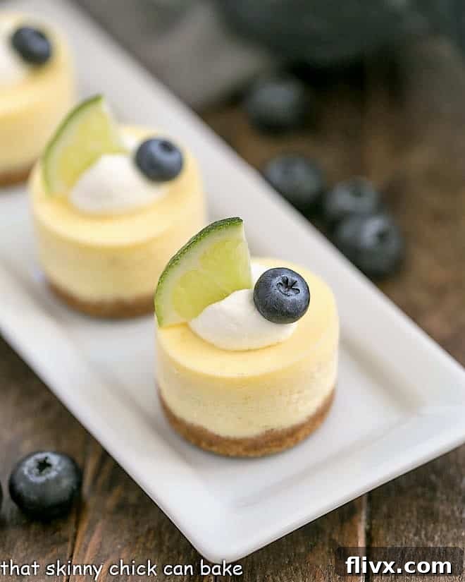 Mini Margarita Cheesecakes topped with cream, a lime slice, and a blueberry on a white tray.