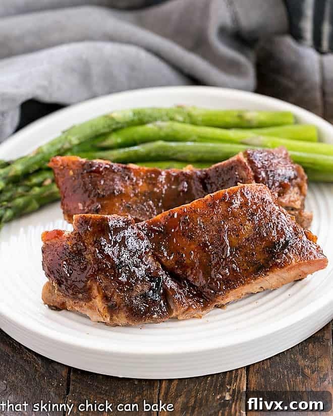 A serving of fall-off-the-bone oven baked ribs on a pristine white plate, accompanied by vibrant green asparagus spears, highlighting the perfect tenderness and presentation of the dish.