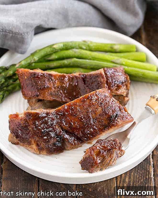A perfectly tender bite of rib meat held on a fork, just removed from a plate featuring two glazed ribs and fresh asparagus, emphasizing the 'fall-off-the-bone' quality.