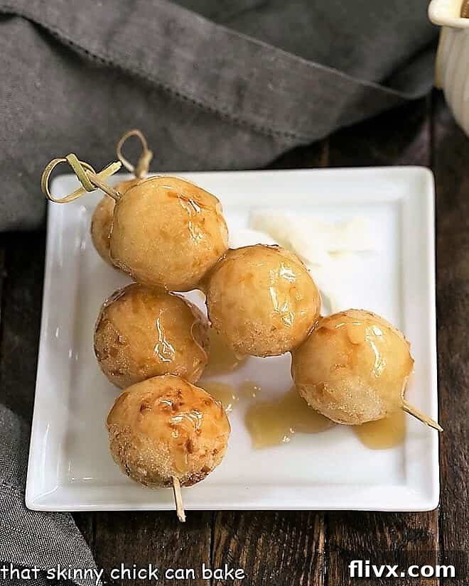 Golden-brown Carioca (Fried Sticky Rice Balls) artfully threaded onto wooden skewers, displayed on a pristine square white plate, highlighting their appealing texture and serving versatility.