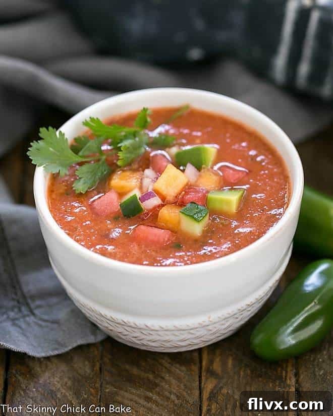 A close-up of vibrant red Watermelon Gazpacho served in a white ceramic bowl, garnished with sliced jalapeños.