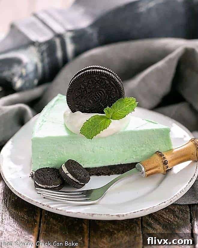 Minty, Green, Frozen No-Bake Dessert with an Oreo Crust: A Delight for Mint Lovers! A perfectly portioned slice of Frozen Grasshopper Pie with an Oreo Crust, beautifully arranged on a white dessert plate, accompanied by extra Oreos and a stylish bamboo-handled fork.